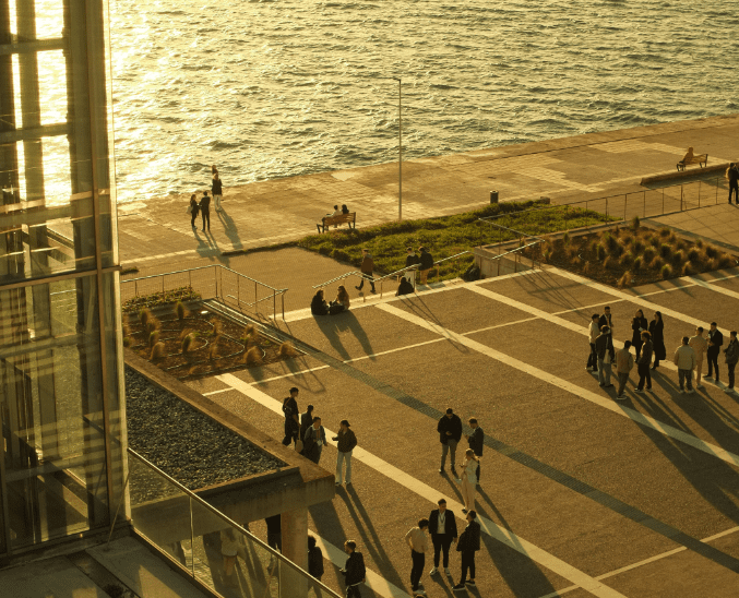 People walking and relaxing on the waterfront promenade near Megaro Mousikis Thessalonikis, with golden sunlight reflecting off the sea.