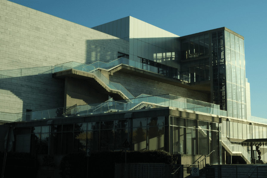 Close-up of the modern white and glass M2 building of Megaro Mousikis Thessalonikis, featuring geometric lines, glass railings, and an external staircase in the sunlight.