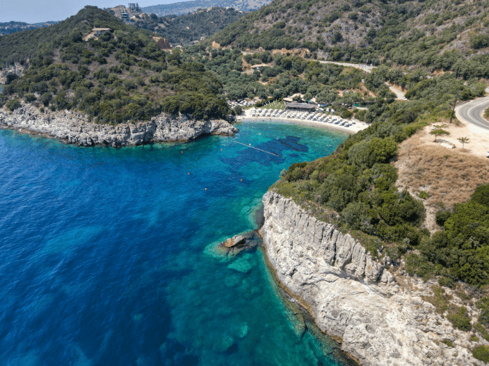 Aerial view of Mikri Ammos Beach in Sivota, surrounded by lush green hills and cliffs, with crystal-clear turquoise waters, sunbeds, umbrellas, and beachside facilities visible along the shoreline.