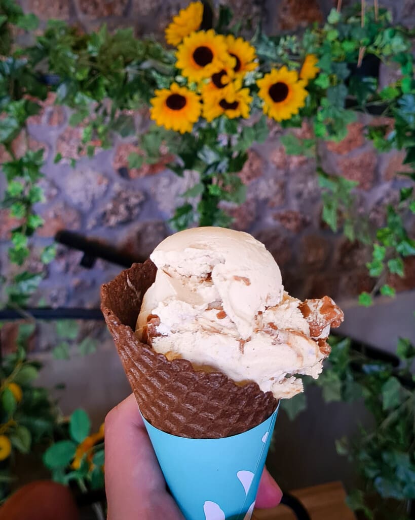 Hand holding a waffle cone with creamy gelato in Litochoro, with sunflowers and green plants in the background.