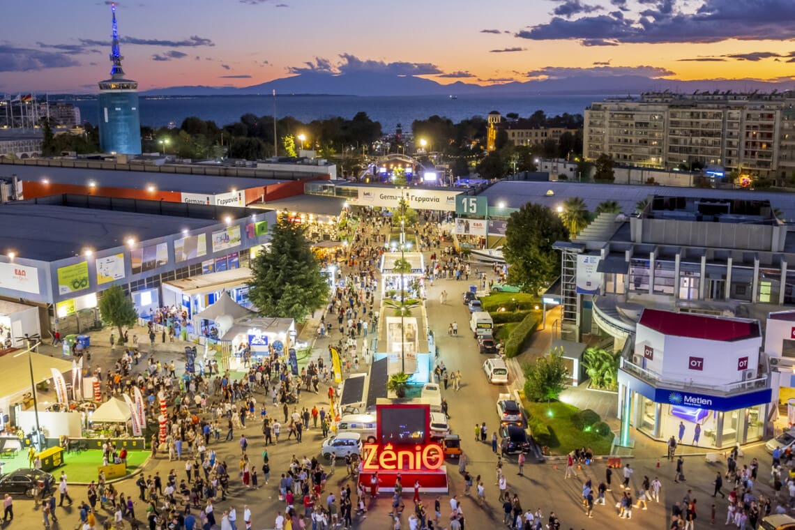 Aerial view of Thessaloniki International Fair with exhibitions, crowds, and cityscape at sunset.